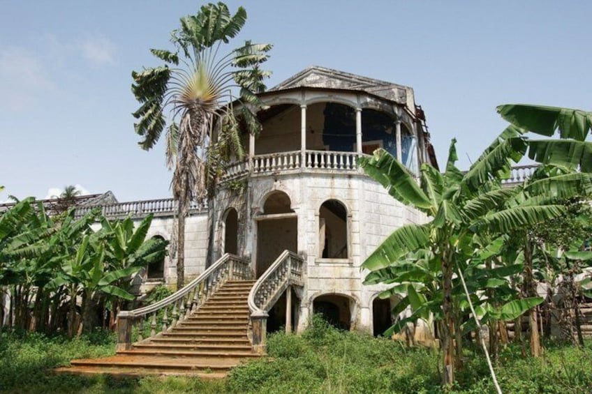 A large, dilapidated building with a staircase leading to a balcony, surrounded by lush greenery and banana trees. 2