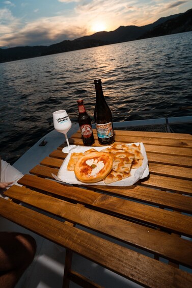 A wooden table with a bottle of beer, a glass, and a plate of food by a lake at sunset. 2