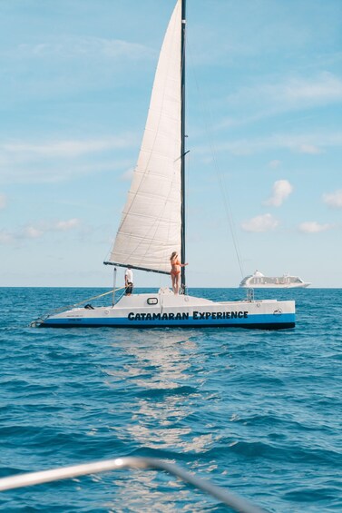 A sailboat with a woman standing at the stern, a large ship in the background, and calm blue waters. 4