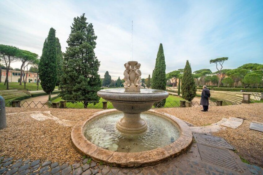 A stone fountain with a statue, surrounded by a gravel path and trees in a park. 5