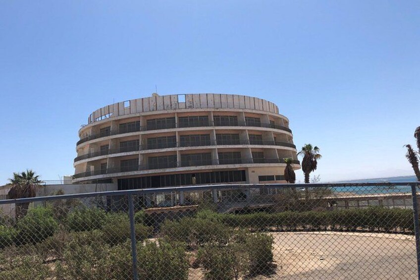 A circular building with multiple balconies, surrounded by a chain-link fence, palm trees, and a beach in the background. 2