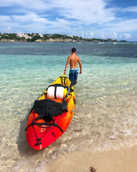 A man walking towards a red and yellow kayak on a beach with clear water and a distant shoreline. 3