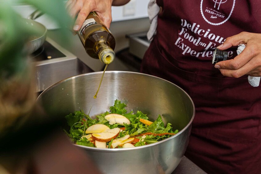 A person pouring olive oil into a salad bowl containing greens and apple slices. 3
