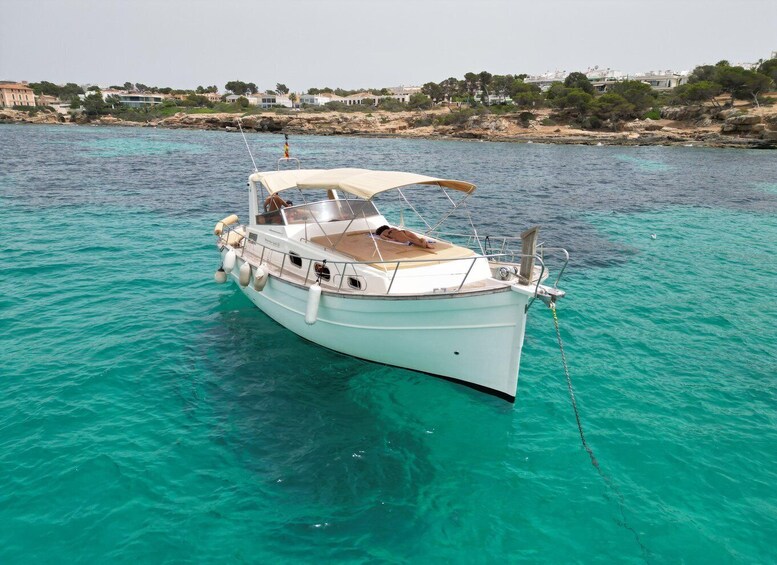 A white boat with a canopy, anchored in clear turquoise water with a rocky shoreline and buildings in the background. 2