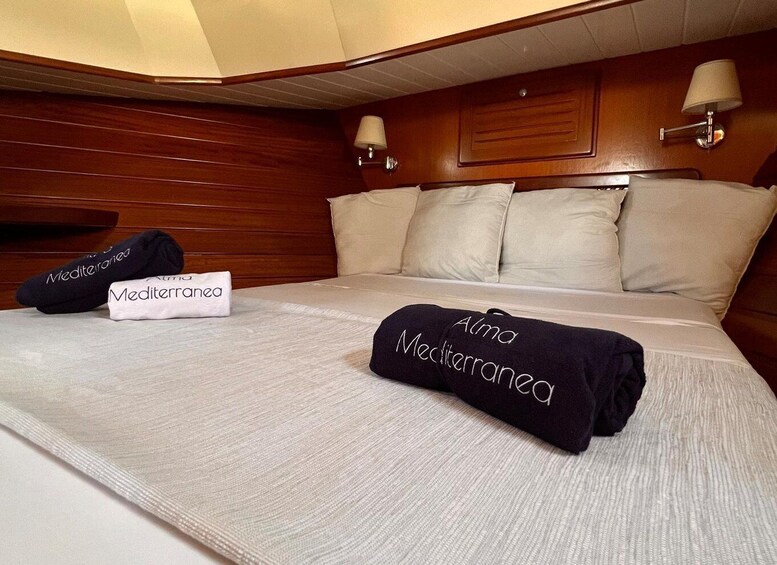A neatly made bed with a white sheet, two pillows, and a folded towel with 'Alma Mediterranea' text. 4