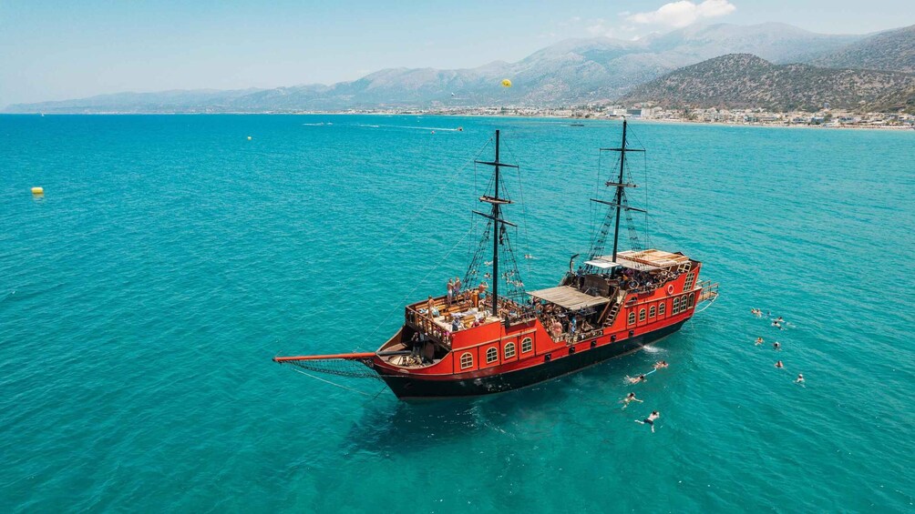 A red ship with multiple masts on turquoise waters, surrounded by a coastal landscape with mountains in the background. 5