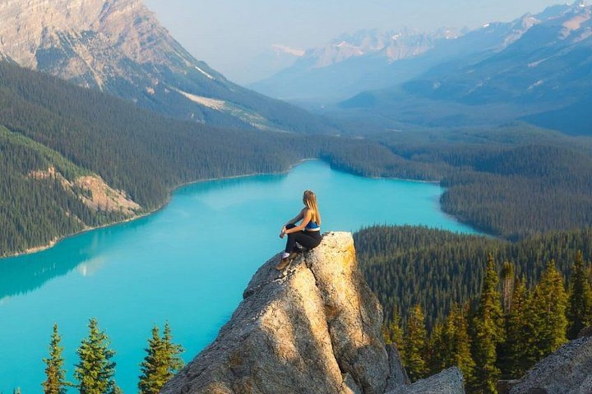 A person sitting on a rock overlooking a turquoise lake surrounded by forested mountains. 1