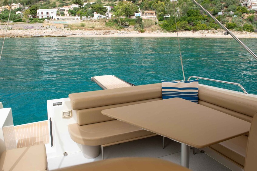 A boat's interior with beige seating and a table, overlooking a turquoise sea and a coastal village. 1