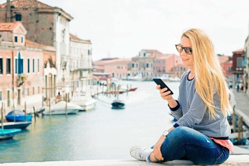 A person sitting by a canal with buildings and boats in the background. 1