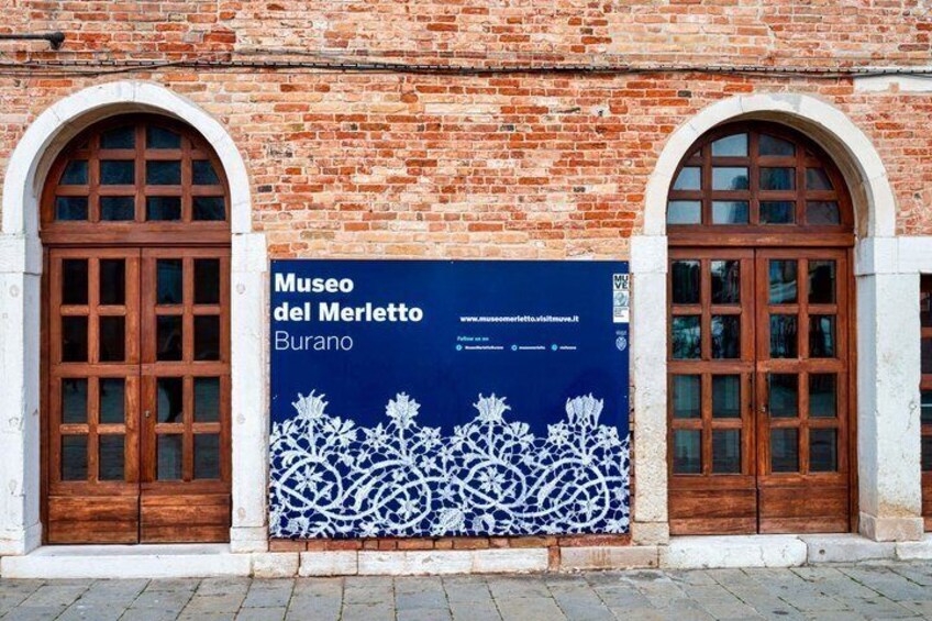 A brick building with two arched wooden doors and a blue sign for Museo del Merletto in Burano. 2