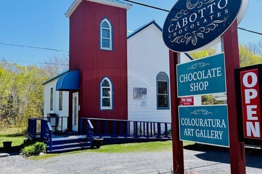 A red building with a blue porch, signs for a chocolate shop and an art gallery, and a sign indicating the shop is for sale. 1