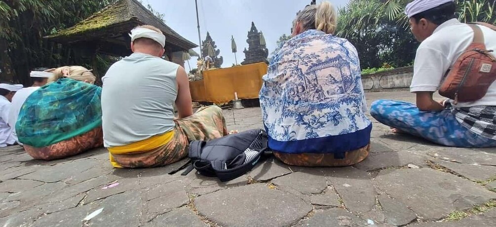 People sitting on the ground, some with bags, in a courtyard with stone flooring and traditional statues in the background. 4