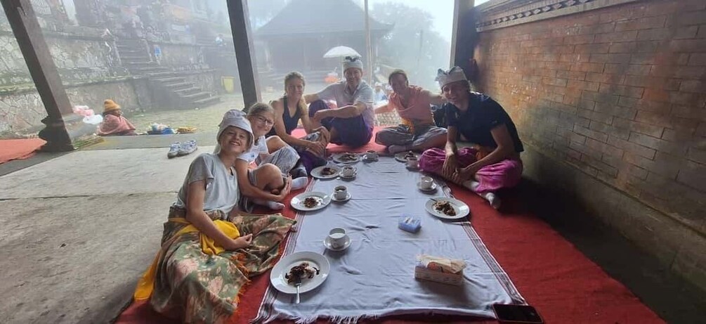 A group of people sitting around a table with food and drinks, outdoors on a red mat. 5