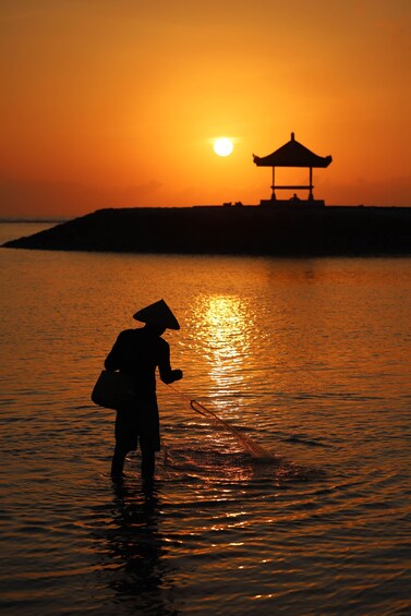 A silhouette of a person in traditional attire standing in shallow water at sunset, with a pagoda in the background. 2