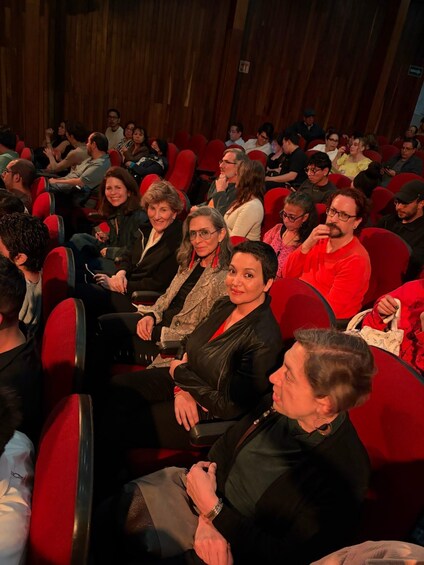 An indoor auditorium with red seats and people seated. 1