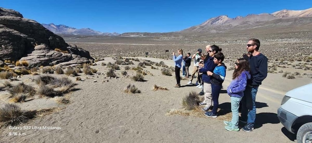 A group of people standing on a desert road with a rocky outcrop and mountains in the background. 3