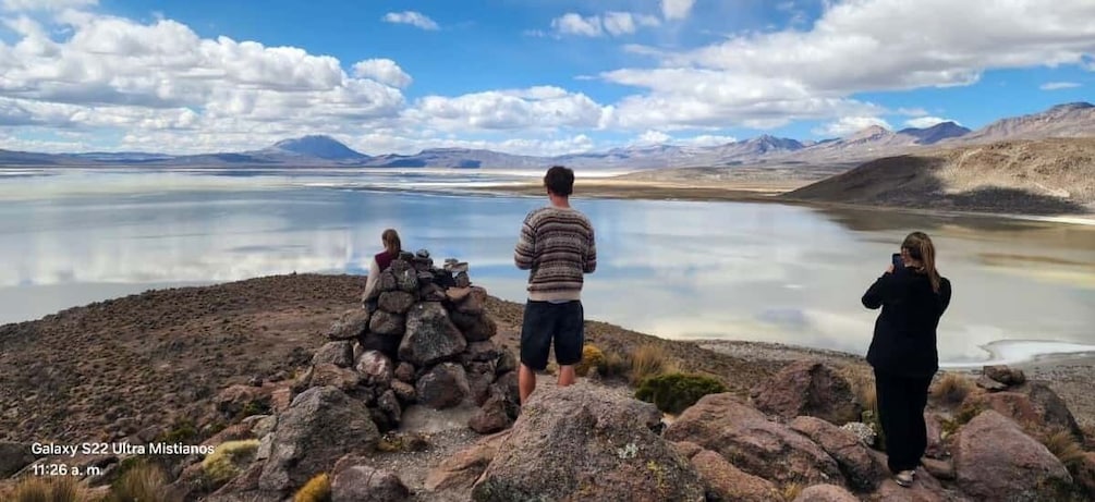 Three people standing on rocky terrain by a calm lake with mountains in the background. 1