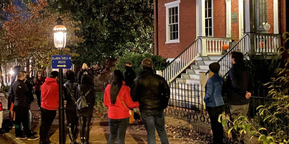 A group of people walking on a sidewalk at night, a street lamp, and a brick building with a staircase. 4