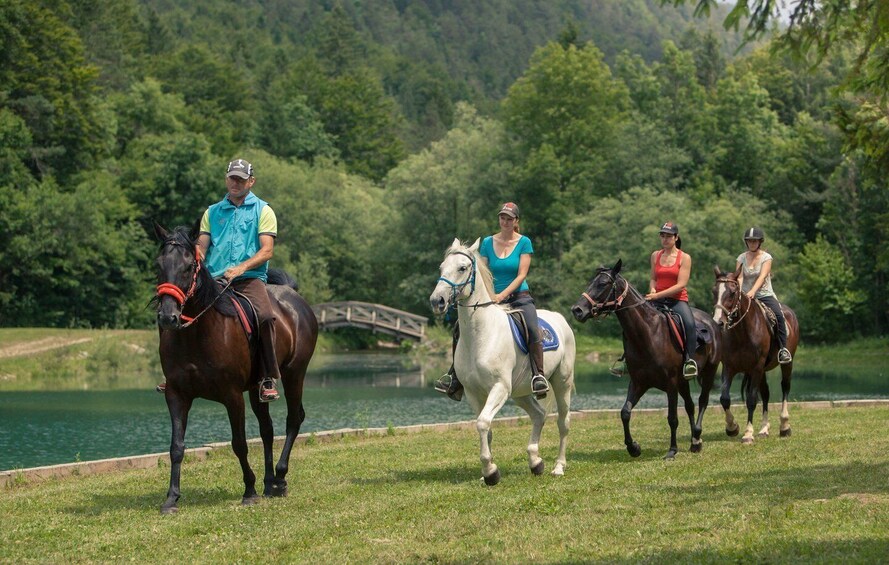Four people riding horses on a grassy field near a body of water with a bridge in the background. 1