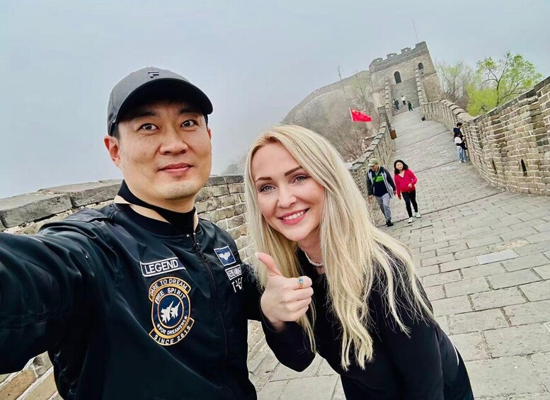 A person in a black jacket with 'LEGEND' and 'FREE SPIRIT' logos, taking a selfie with a historic stone wall and a red flag in the background. 1