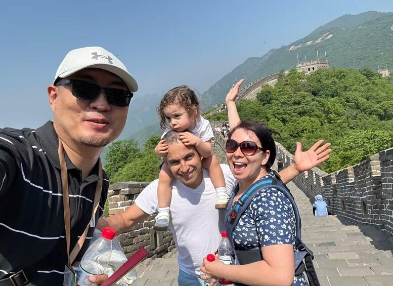 A family on the Great Wall of China with a child on an adult's shoulders, a clear sky, and mountains in the background. 2