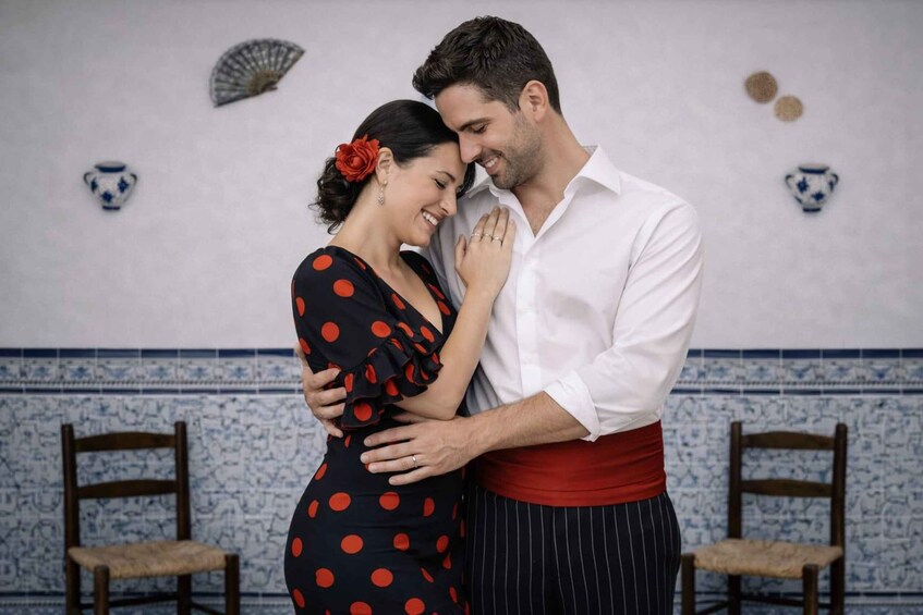 A couple embracing in a room with blue and white patterned tiles, two wooden chairs, and decorative wall pieces. 3