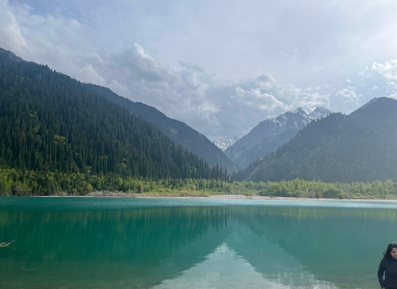 A calm lake surrounded by dense forests and snow-capped mountains under a partly cloudy sky. 2