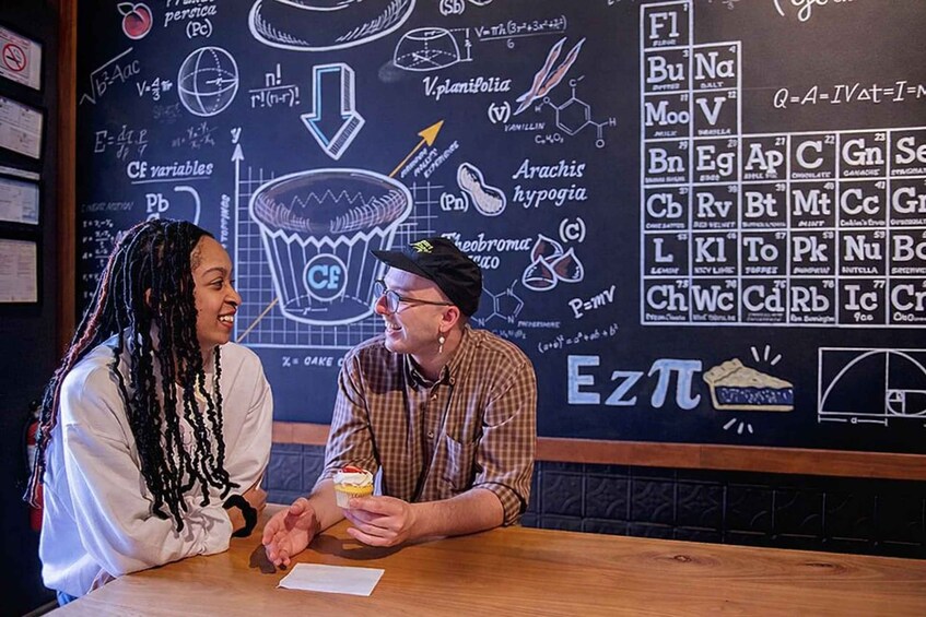Two people at a table with a chalkboard filled with scientific and mathematical equations and symbols. 3