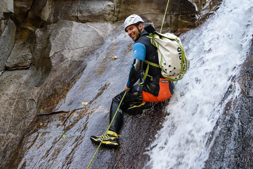 A person rappelling down a wet rock face with a backpack and climbing gear. 1