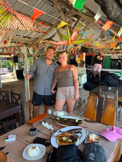 Two people standing at a wooden table with food, under a colorful tent. 2