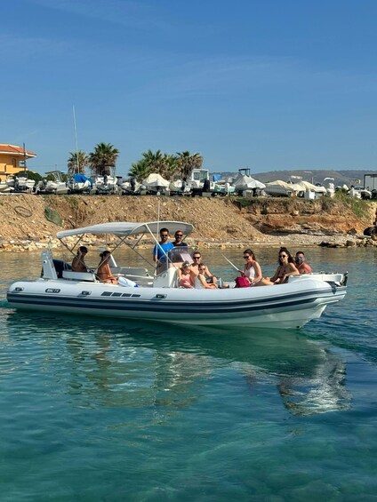 A group of people on a small motorboat on clear blue water, with a beach and parked vehicles in the background. 1