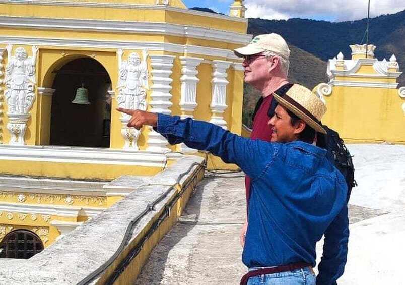 A person pointing at a yellow building with white statues and a bell. 1
