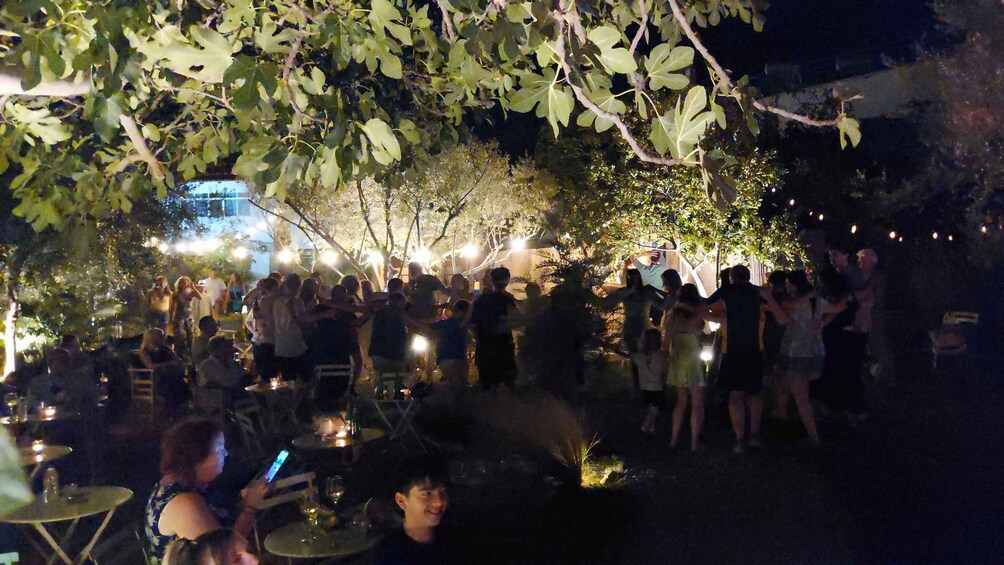 A nighttime outdoor gathering with people under trees, tables with drinks, and ambient lighting. 3