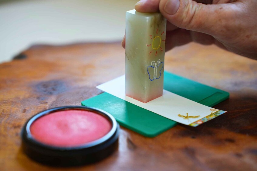 A hand holding a cylindrical object with a sun and butterfly drawing, a green envelope, and a red container on a wooden surface. 3