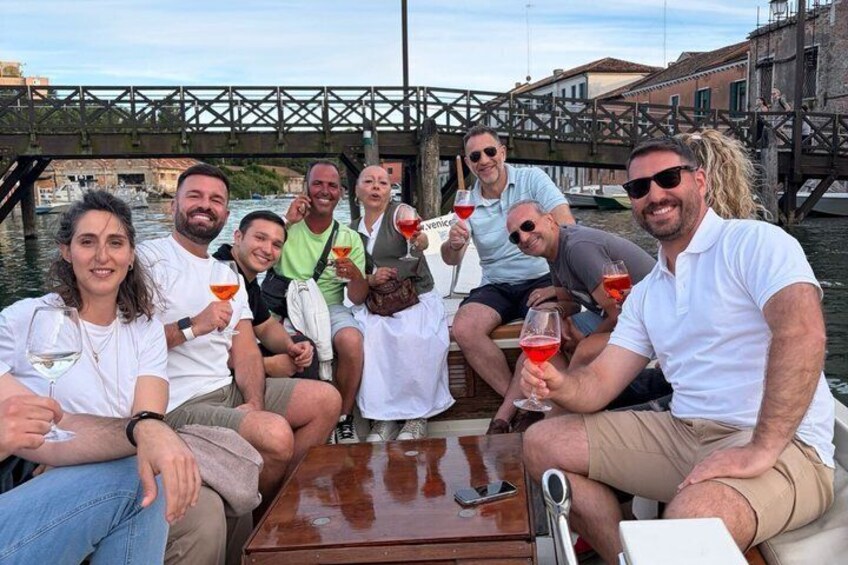 A group of people on a boat with drinks, a wooden table, and a bridge in the background. 2