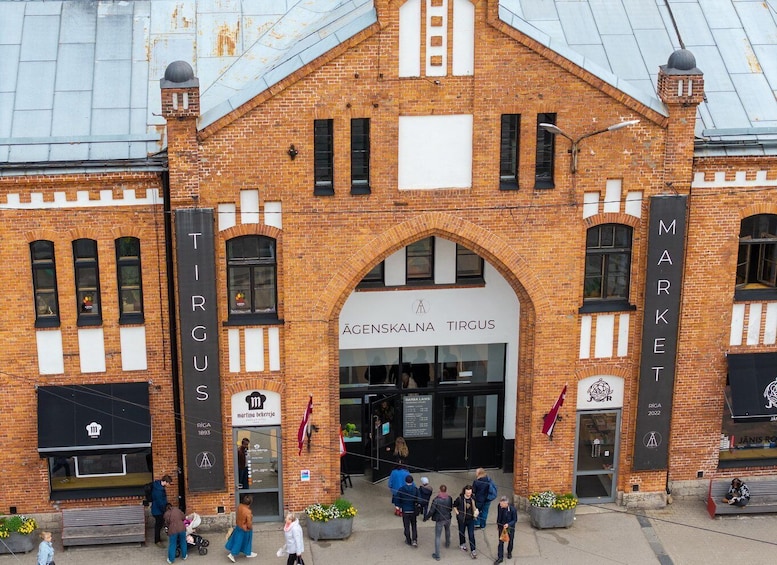 A brick building with 'TIRGUS' and 'MARKET' signs, a storefront with 'AGENSKALNA TIRGUS' and 'MARTINO BECKERJA 1893' signs, and people walking in front. 1