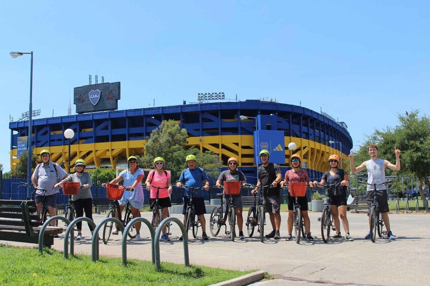 A group of people on bicycles in front of a stadium with a blue and yellow color scheme. 3