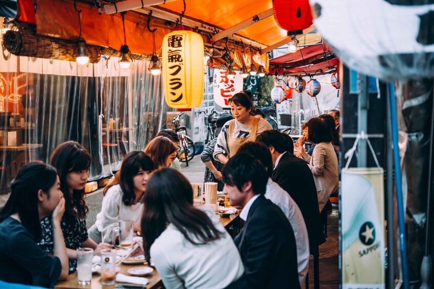 A group of people dining at an outdoor restaurant with hanging lanterns and a sign indicating a ramen stall. 2