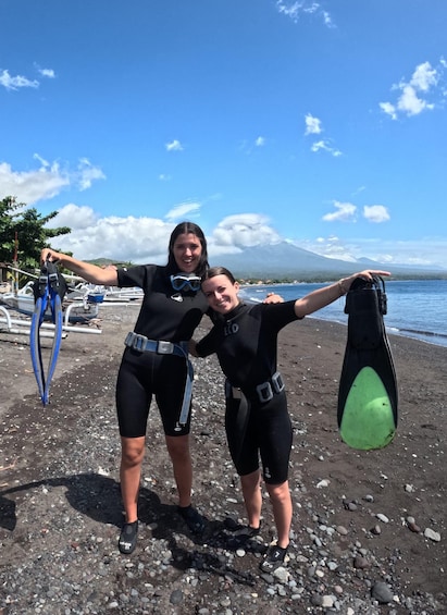 Two people in wetsuits holding diving fins on a rocky beach with a mountain in the background. 2
