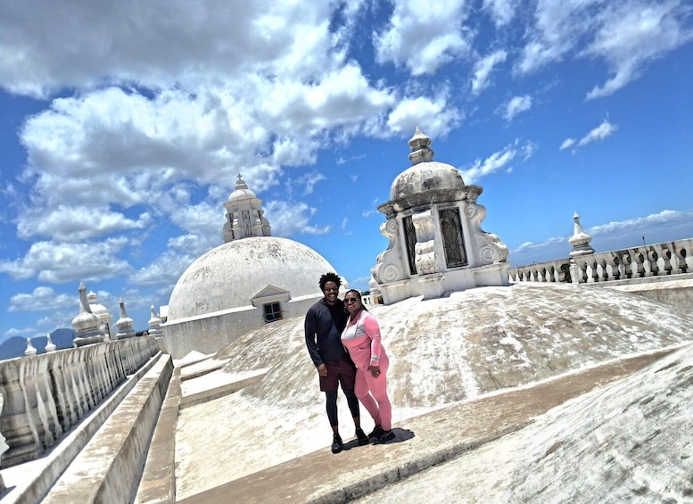 A couple standing on a stone staircase with a large white dome and smaller domes in the background under a partly cloudy sky. 5
