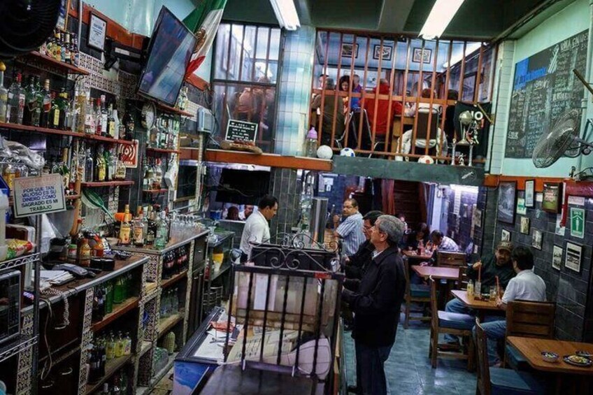 A crowded bar with shelves of bottles, a man standing, and a balcony with people. 1
