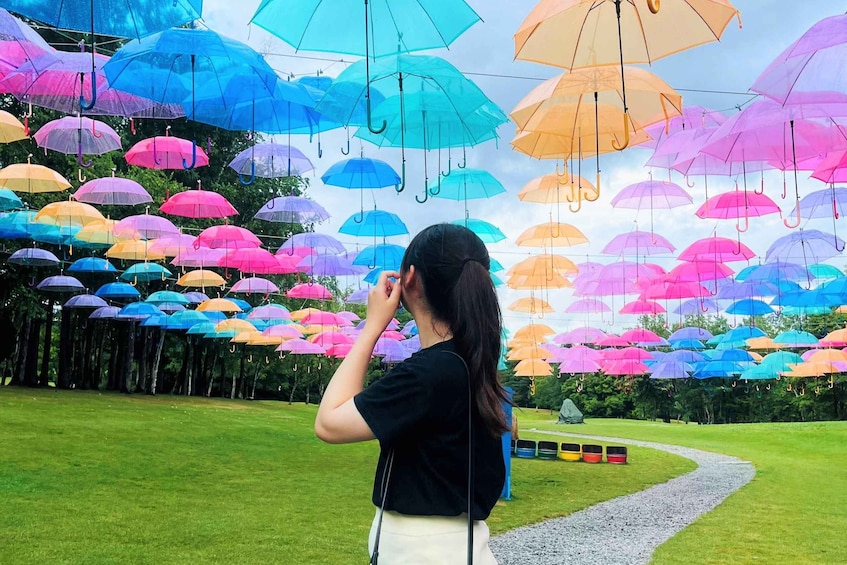 A person standing in a park with a path, surrounded by numerous colorful umbrellas suspended from above. 2