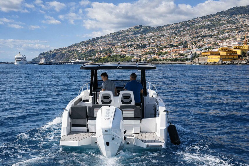 A speedboat with two passengers on a clear day, heading towards a coastal town with a prominent yellow building. 3