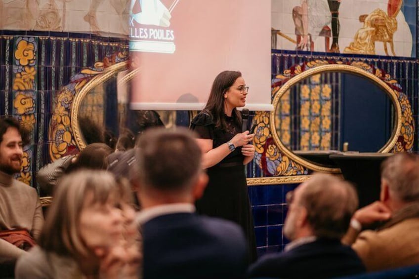 A person giving a presentation in front of a crowd with a decorative tiled wall in the background. 1