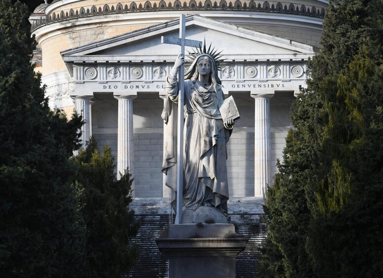 A statue of a robed figure holding a cross, with inscriptions on the building behind it. 3
