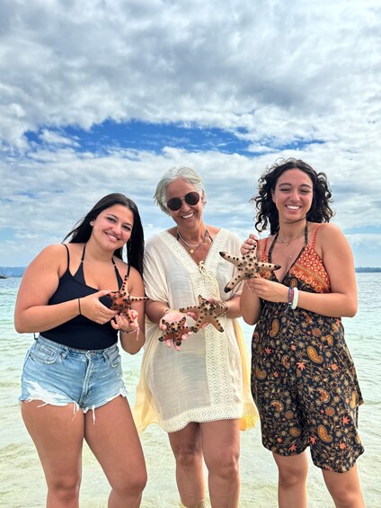 Three people holding starfish on a beach with clear water and a cloudy sky. 3