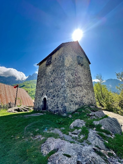 A stone building with a sloped roof, surrounded by grass and rocks, under a clear blue sky with the sun shining. 1