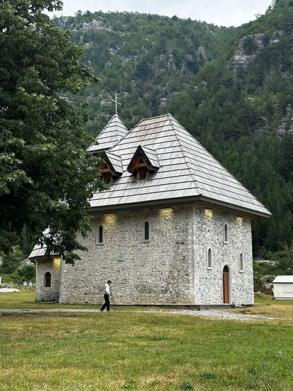 A stone church with a steep roof and a bell tower, surrounded by a grassy field and trees. 2