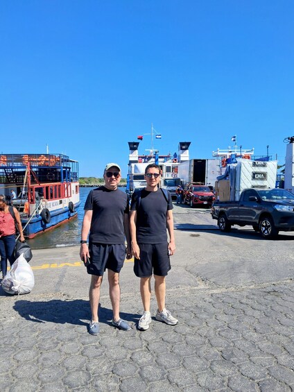 Two people standing in a port with boats and trucks in the background. 4