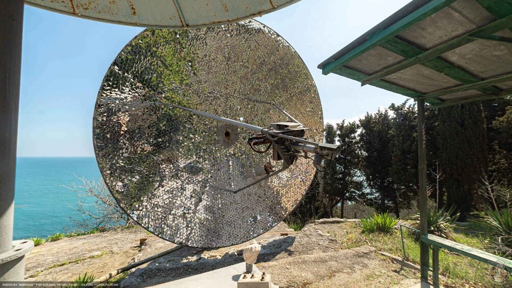 A large satellite dish with a camera mounted on it, overlooking a coastal area with trees and a clear sky. 3